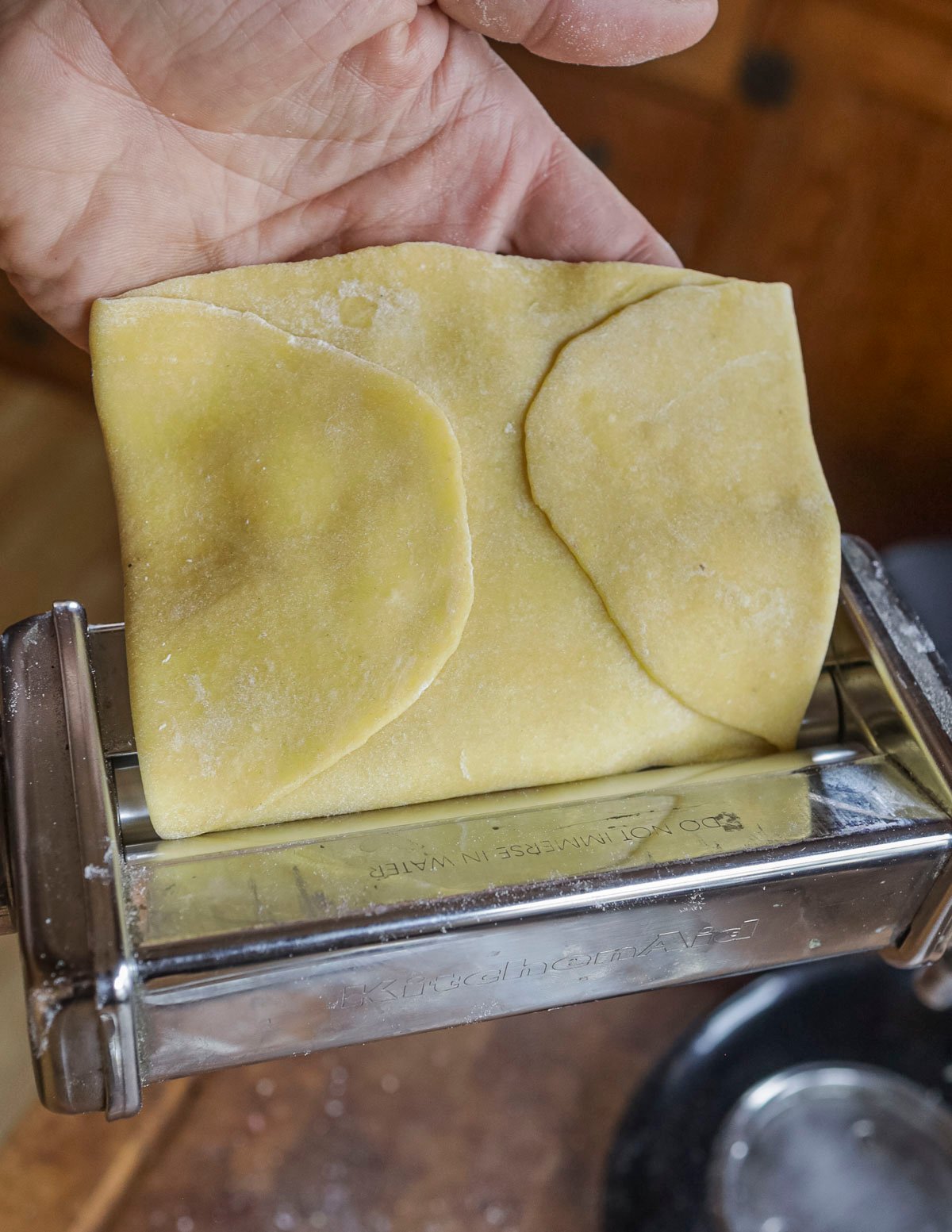 A hand putting pasta dough through a roller illustrating how it should be folded over itself to perfectly fit the width of the pasta roller. 