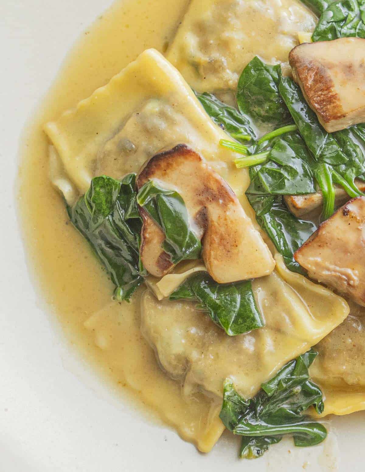 A plate of cooked porcini mushroom ravioli on a plate with a brown butter sauce, spinach and sauteed fresh porcini.