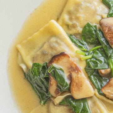 A plate of cooked porcini mushroom ravioli on a plate with a brown butter sauce, spinach and sauteed fresh porcini.