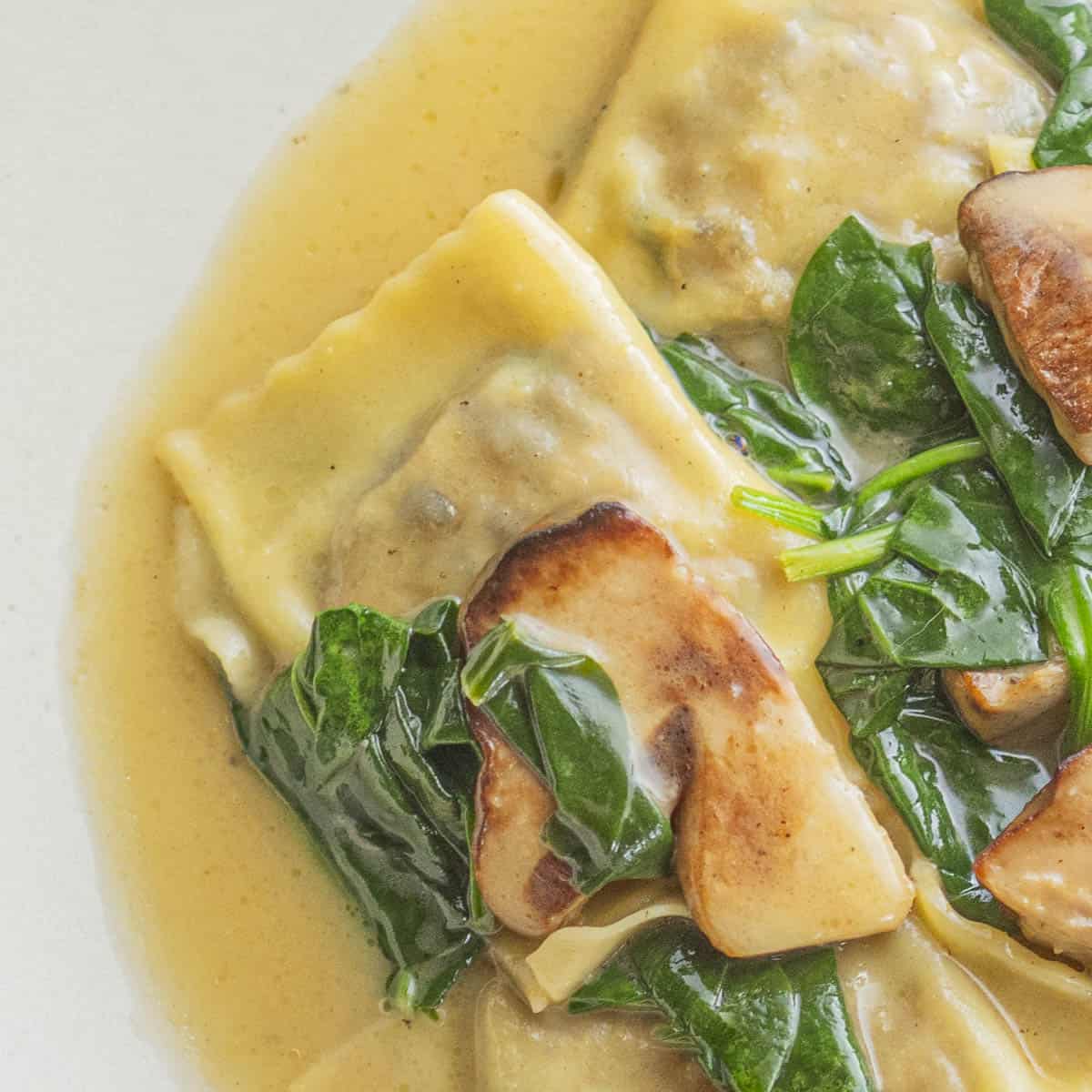 A plate of cooked porcini mushroom ravioli on a plate with a brown butter sauce, spinach and sauteed fresh porcini.