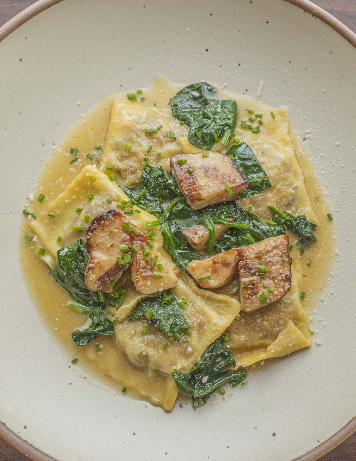 A plate of cooked porcini mushroom ravioli on a plate with a brown butter sauce, spinach and sauteed fresh porcini. 