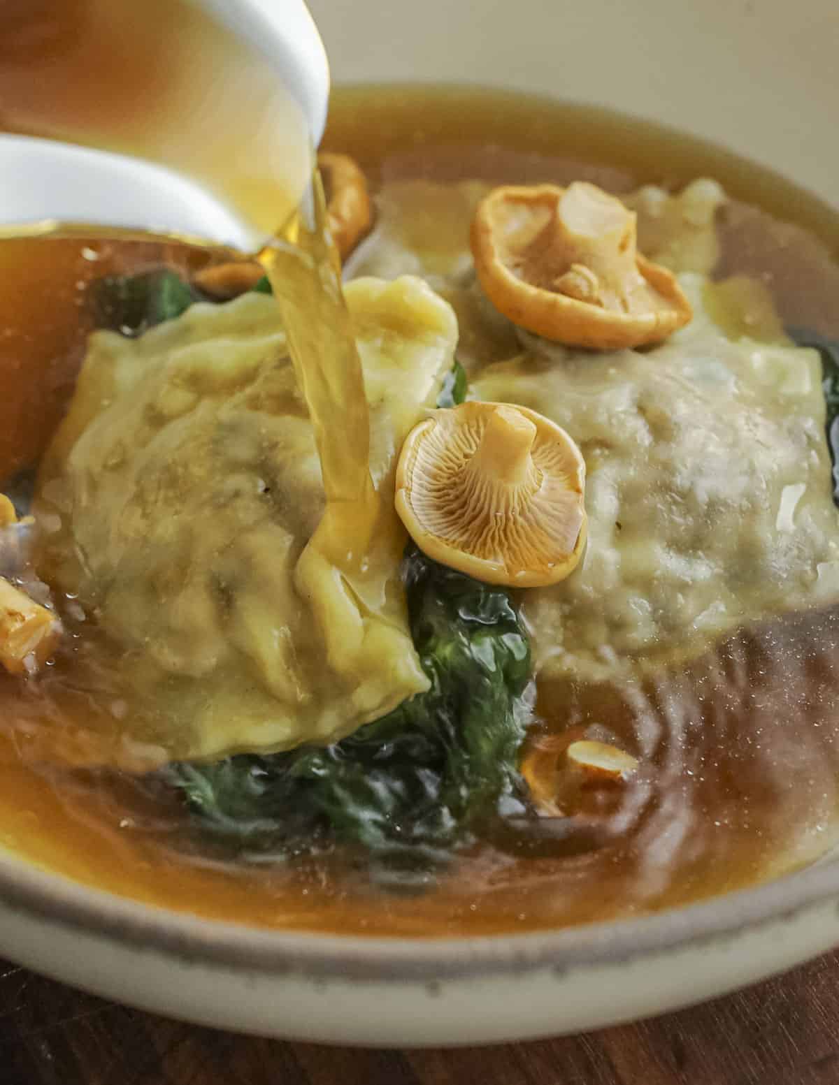 Pouring broth into a bowl of mushroom ravioli with spinach and chanterelle mushrooms. 