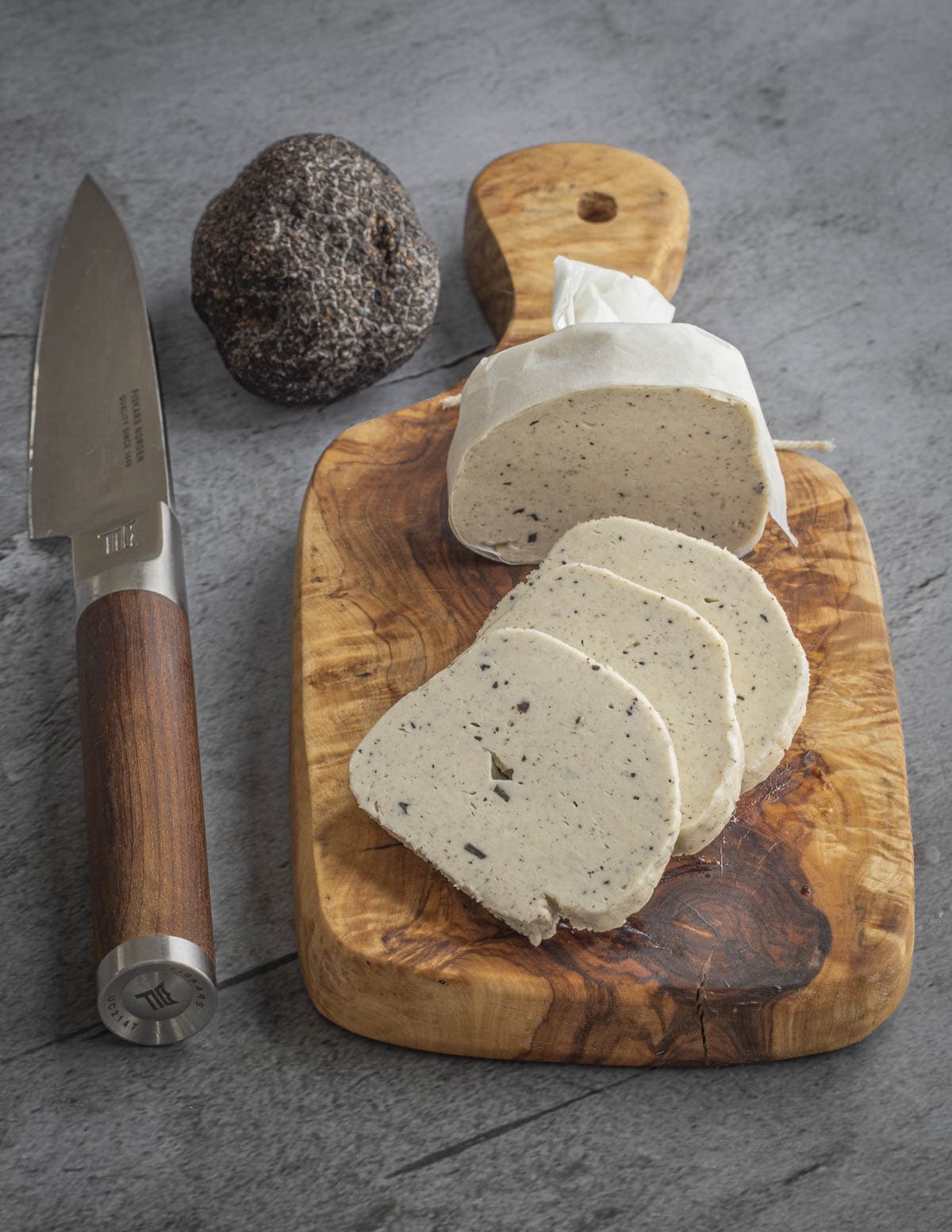 A log of black truffle butter on a small cutting board next to a knife and a whole Tuber melanosporum. a