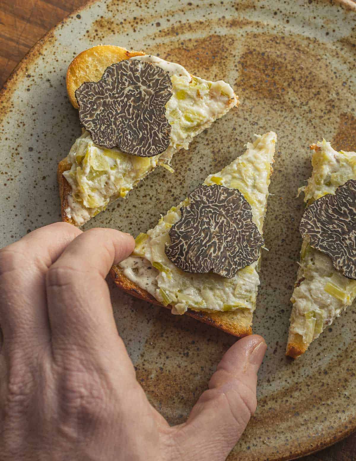 A hand reaching to grab a slice of toast with leeks and fresh sliced black truffles.