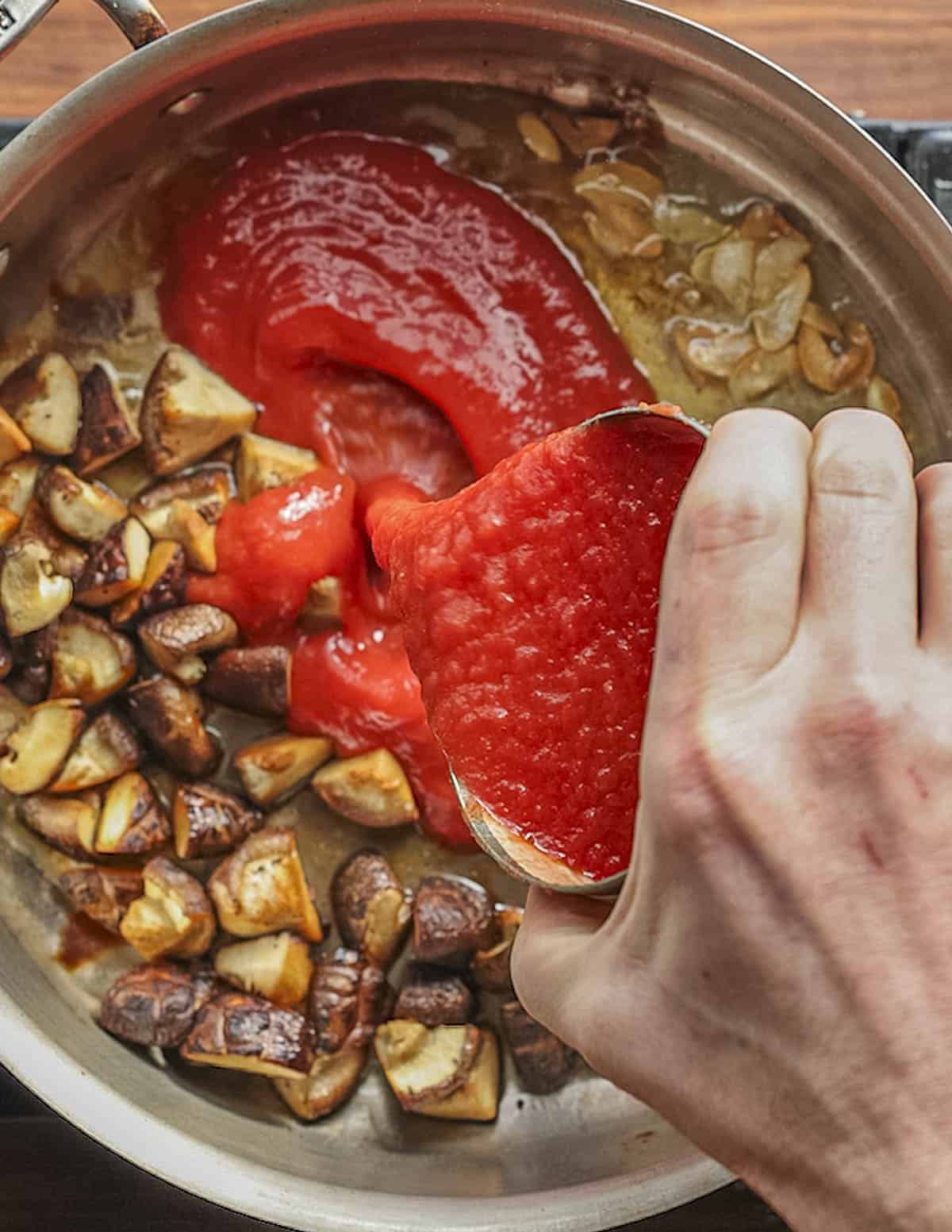 Adding tomato puree to a pan of cooked mushrooms and garlic. 