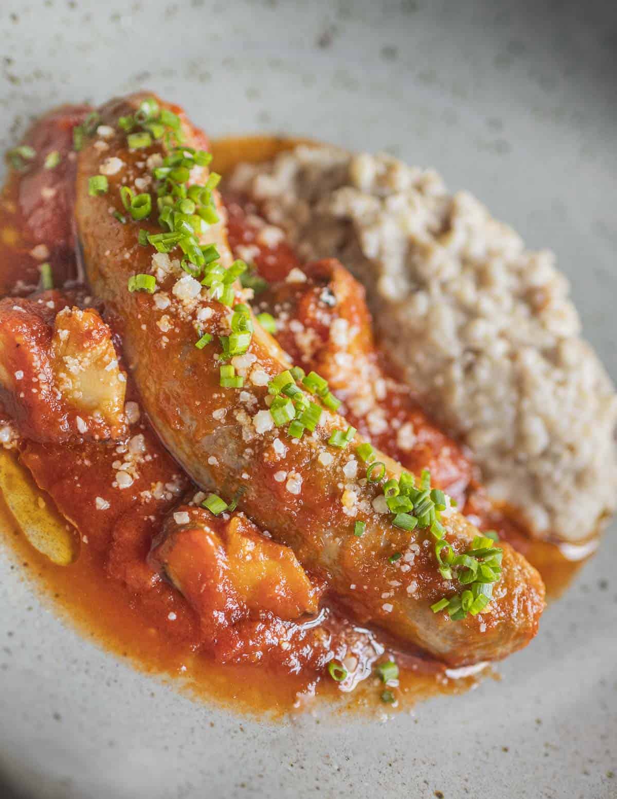 A baked Italian sausage on a plate with tomato sauce, mushrooms parmesan cheese and polenta. 