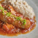 A baked Italian sausage on a plate with tomato sauce, mushrooms parmesan cheese and polenta.