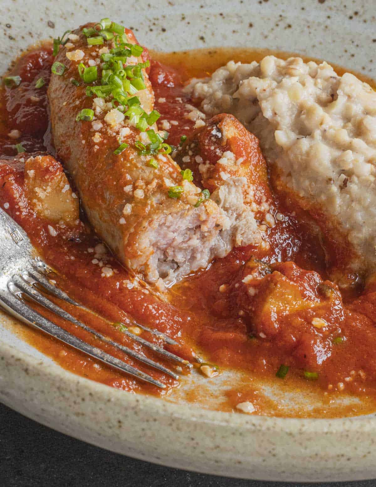 A fork cutting into a sausage with tomato sauce and mushrooms served with polenta. 