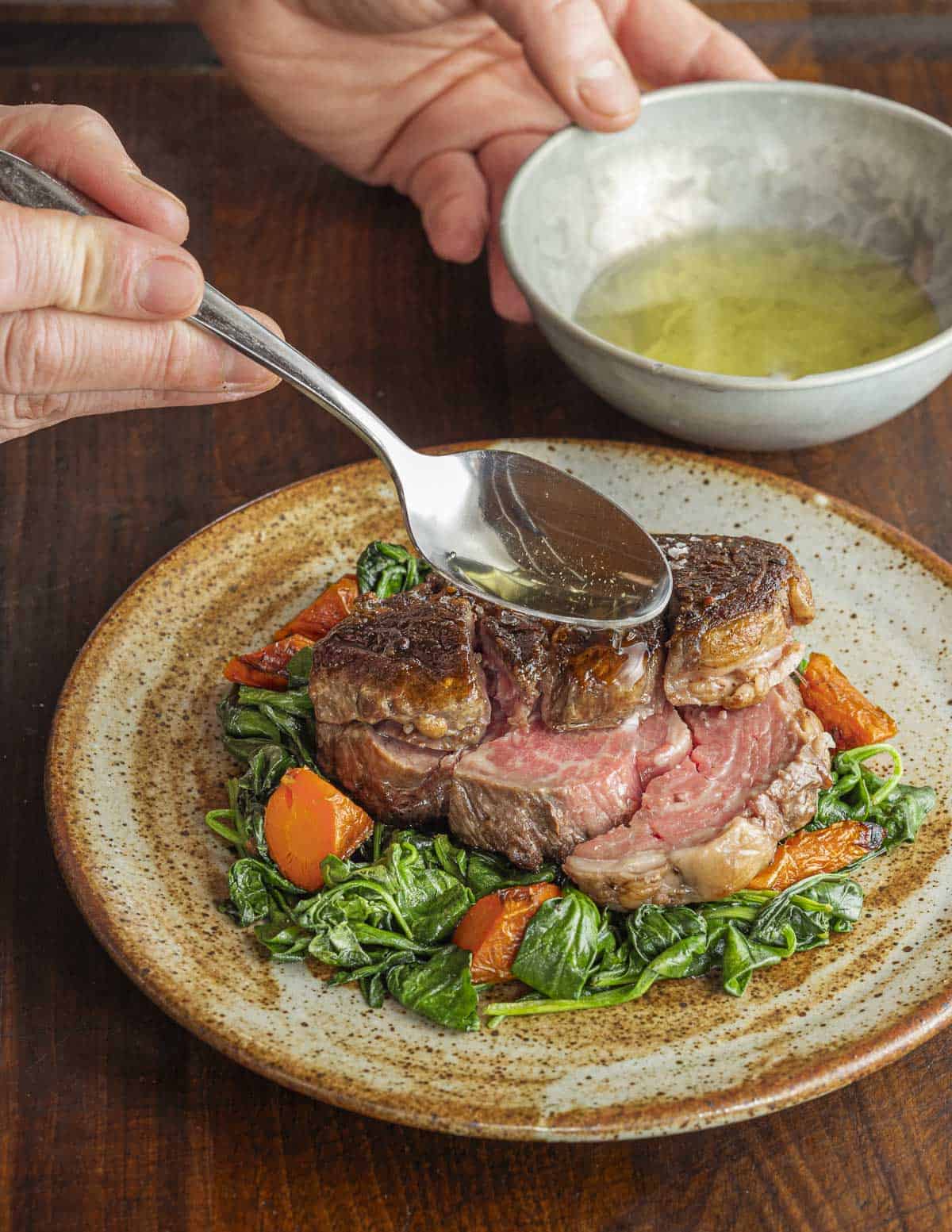 Spooning lemon oil over a cooked deckle steak on top of cooked spinach and carrots. 