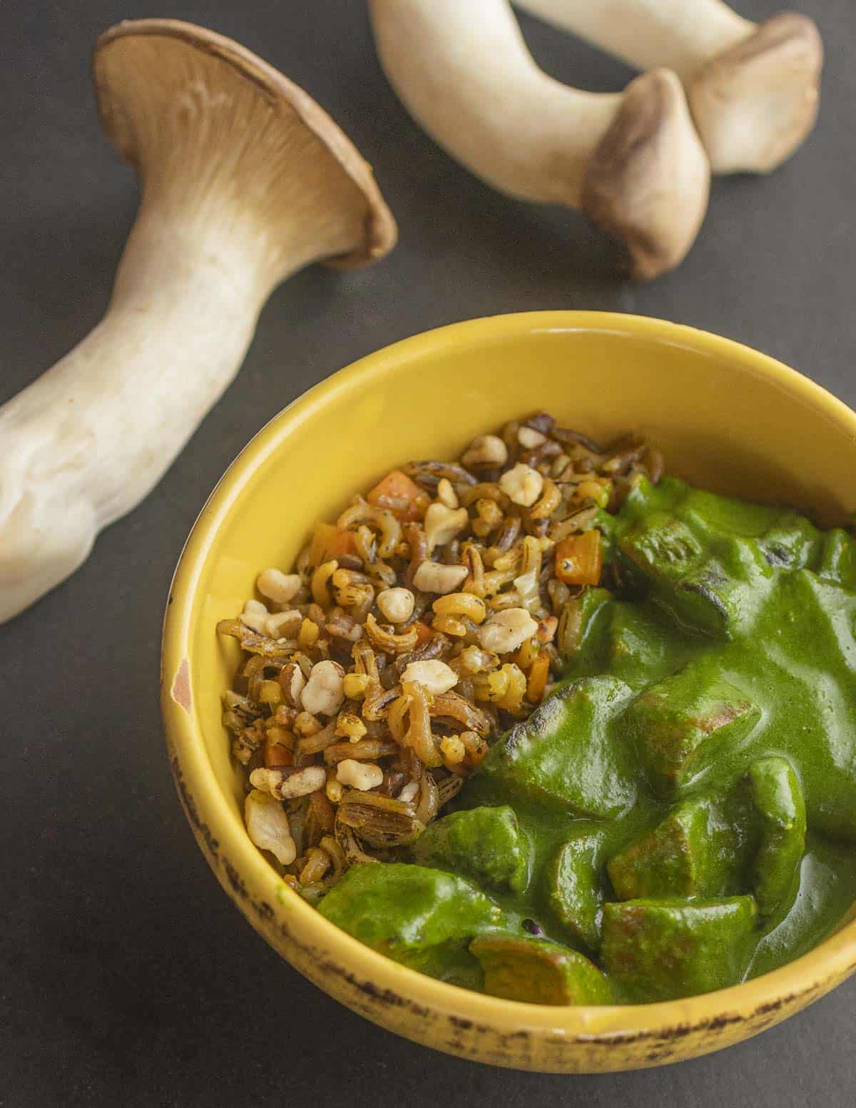 A bowl of mushroom saag curry with wild rice and king trumpet mushrooms in the background. 