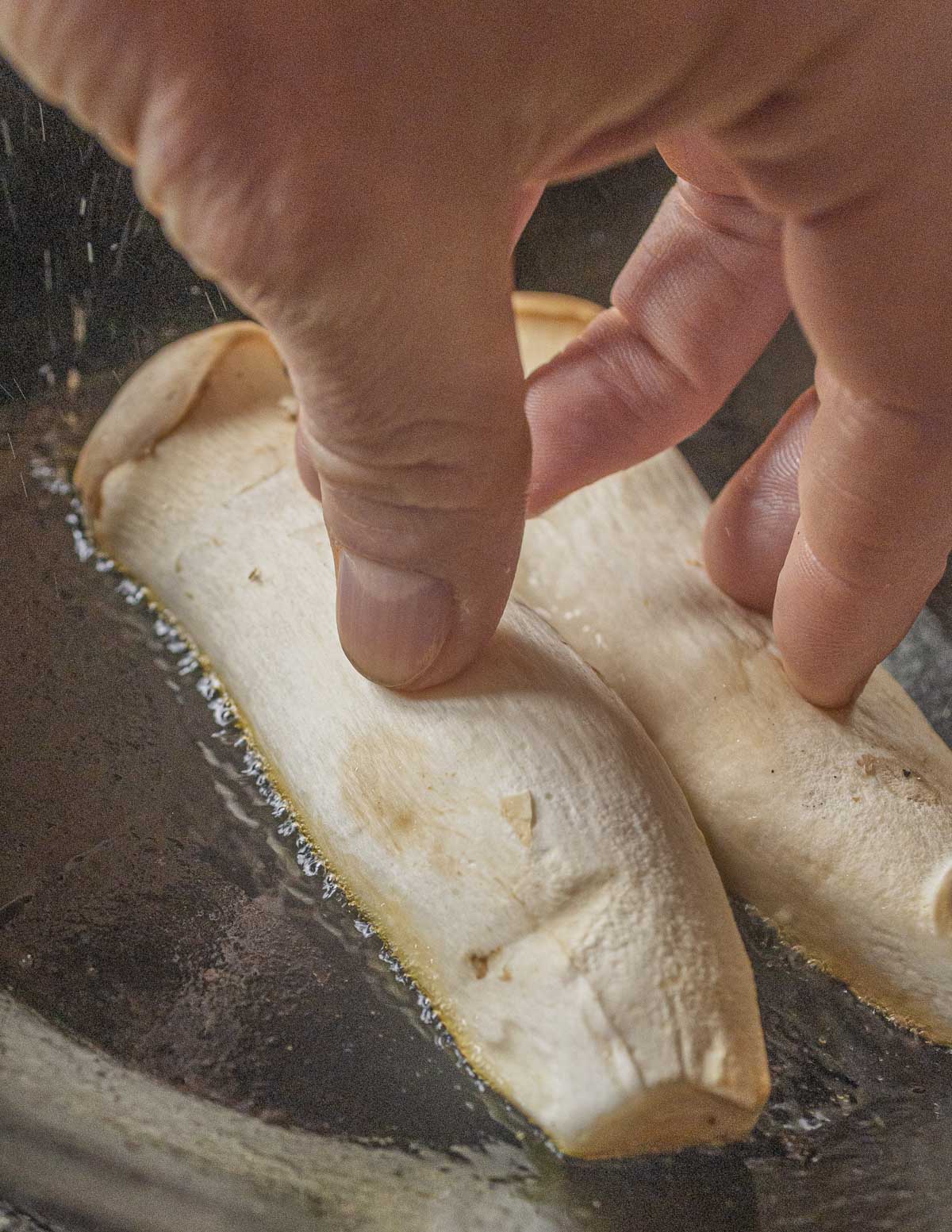 A hand pressing down on two cut king oyster mushrooms to help them brown in a pan. 