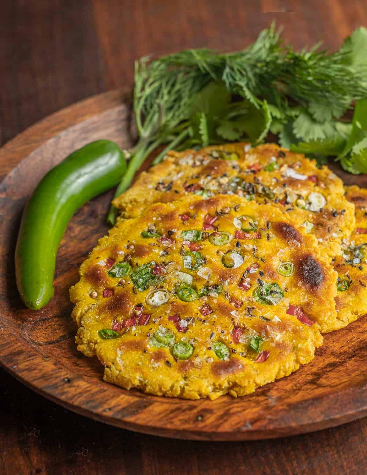 Missi roti or Indian chickpea flatbread on a wooden plate next to a green chili and cilantro. 