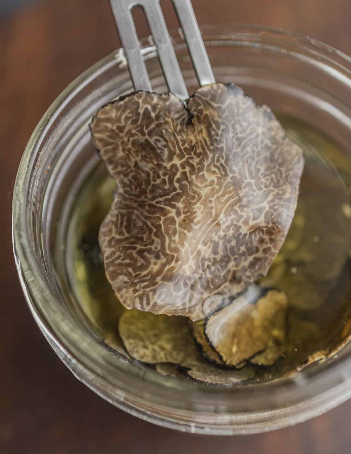 A fork removing a slice of truffles from a jar of black truffle slices in oil.