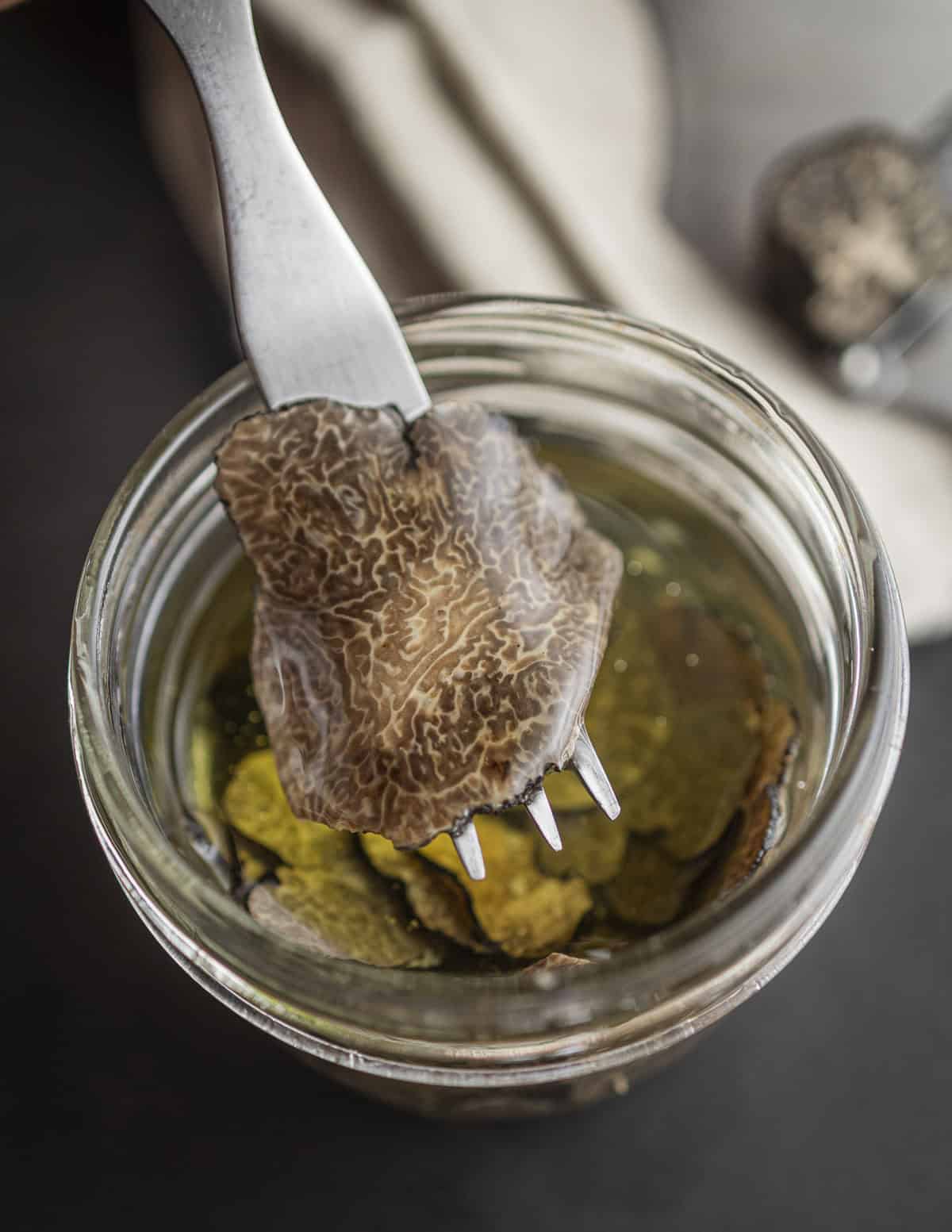 A fork removing a slice of truffles from a jar of black truffle slices in oil.