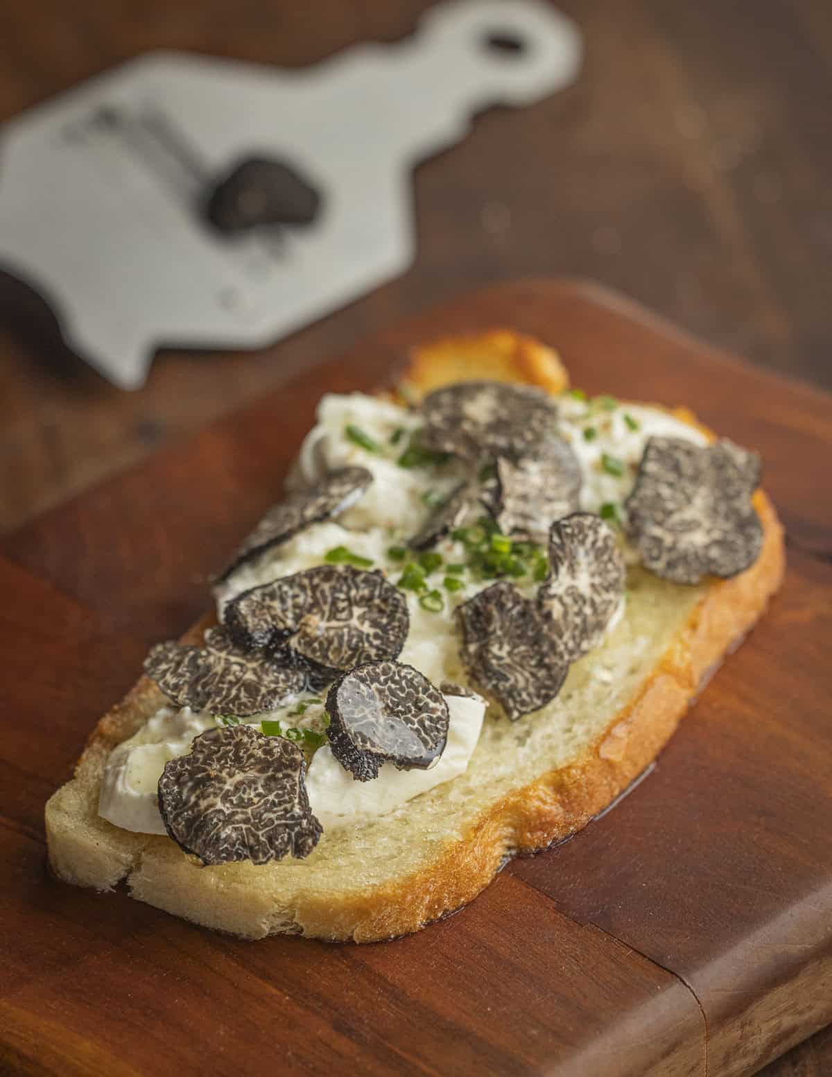 A slice of toast with burrata cheese, black truffles and black truffle oil with a truffle slicer and truffles in the background. 