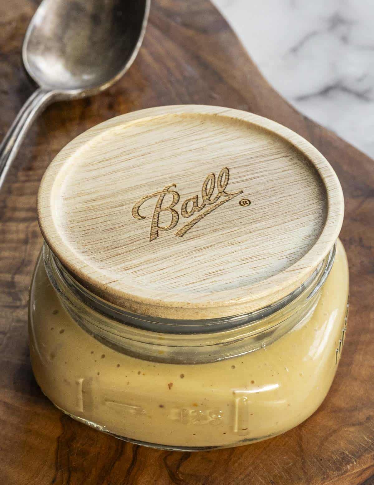 A jar of  dijon maple vinaigrette next to a spoon on a cutting board. 
