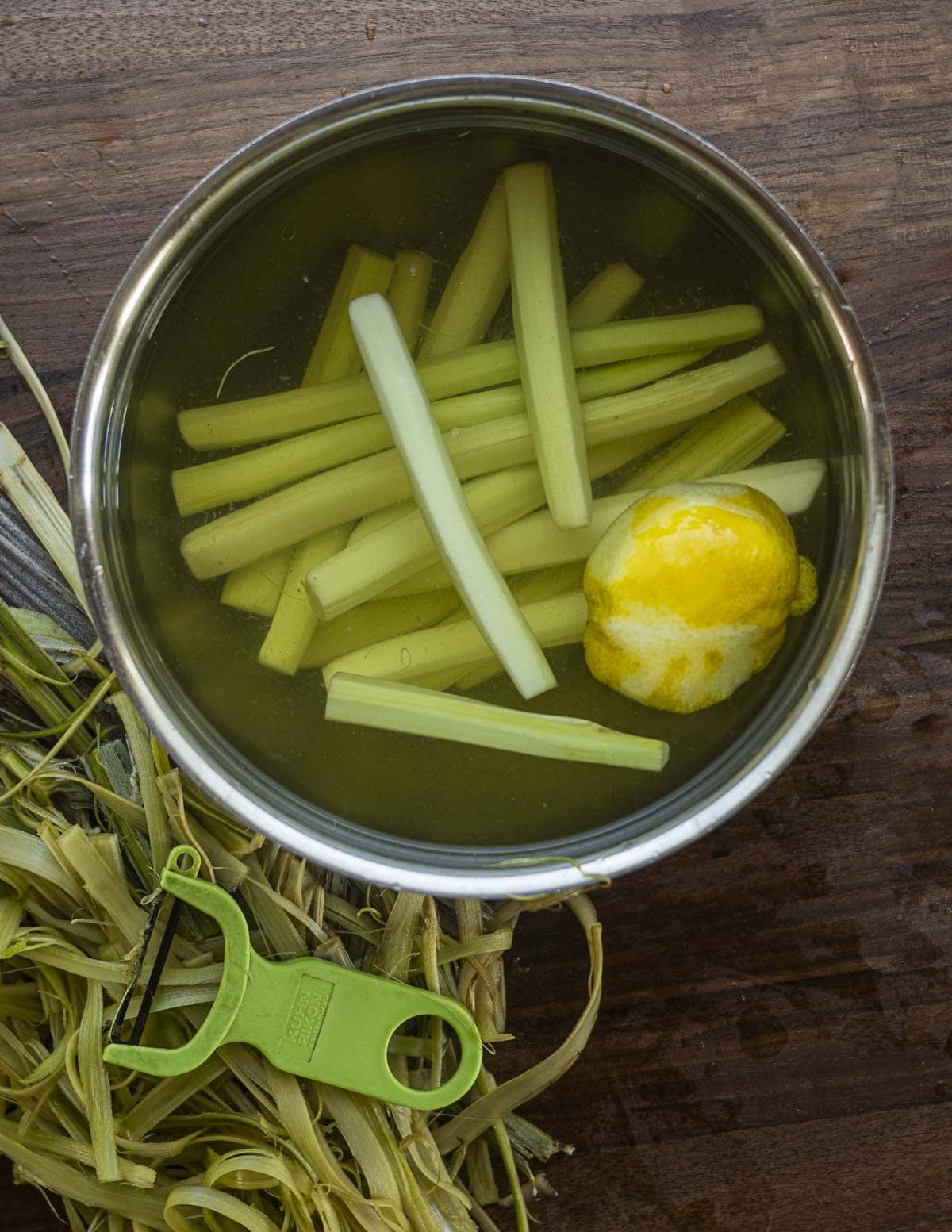 A bowl of edible burdock flower stalks being held in acidulated water to prevent discoloration next to a vegetable peeler. 