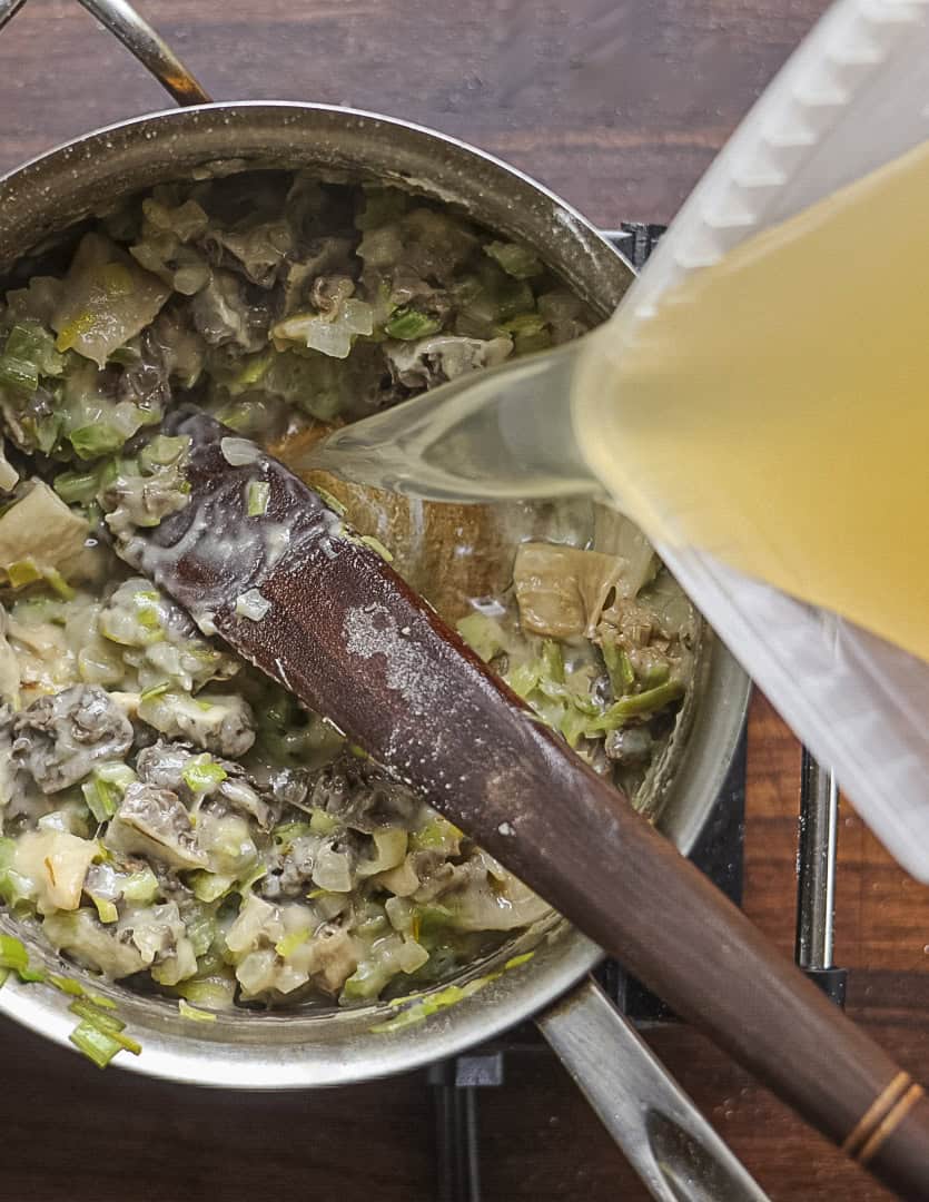 Gradually adding chicken stock to a pan of morel mushroom soup. 