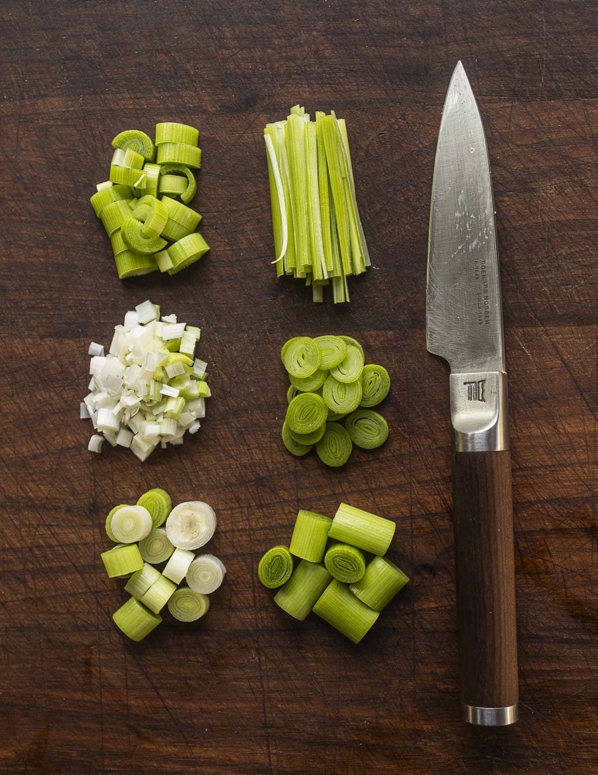 Green garlic cut into 6 different shapes next to a paring knife on a cutting board. 