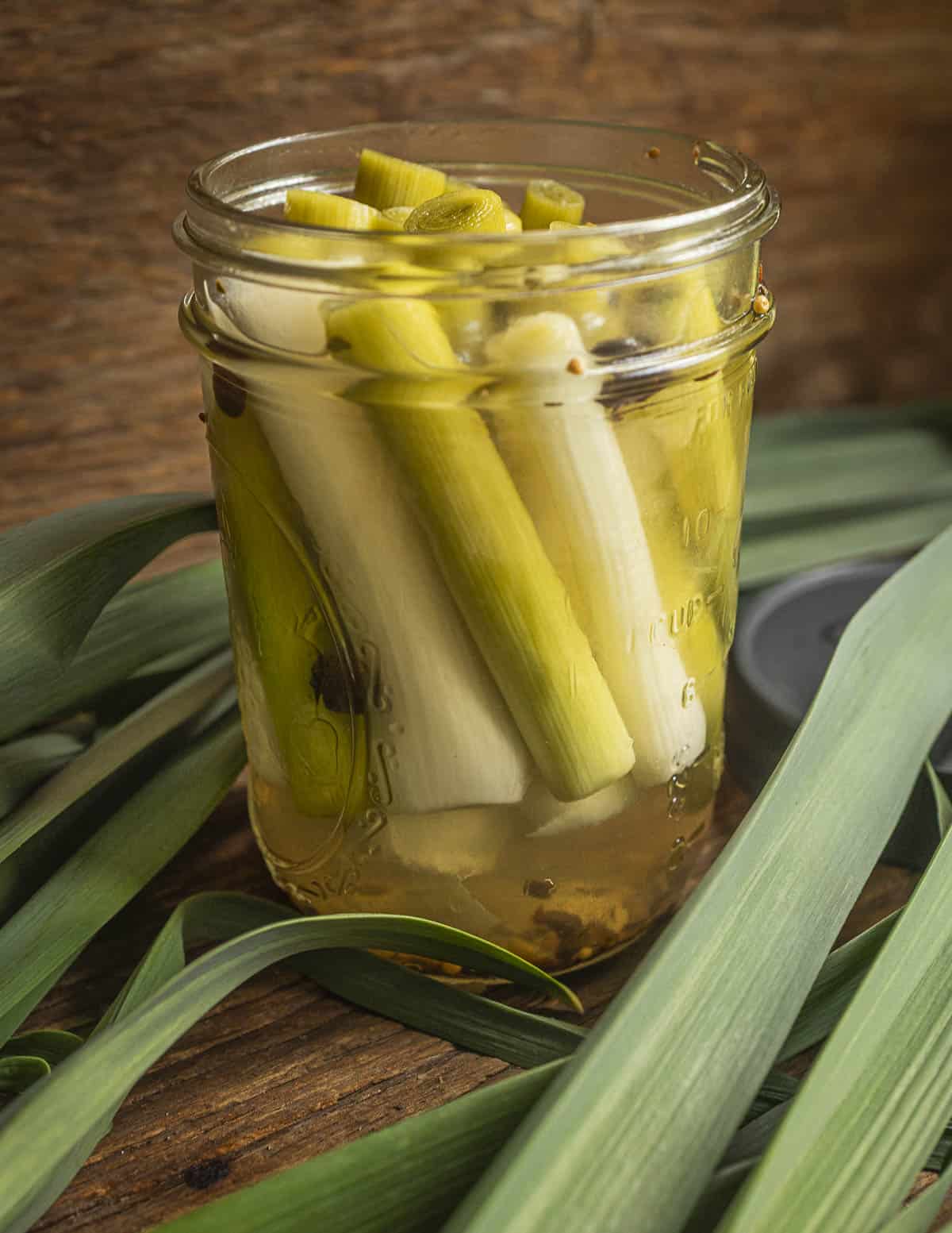 A jar of pickled green garlic stems surrounded by green garlic leaves.