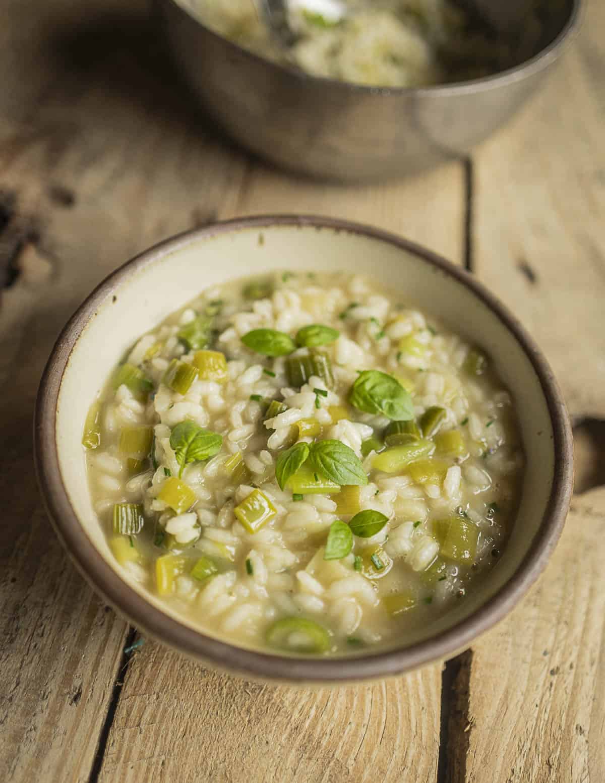 A green garlic risotto in a bowl next to a pan of risotto garnished with fresh sprigs of basil. 