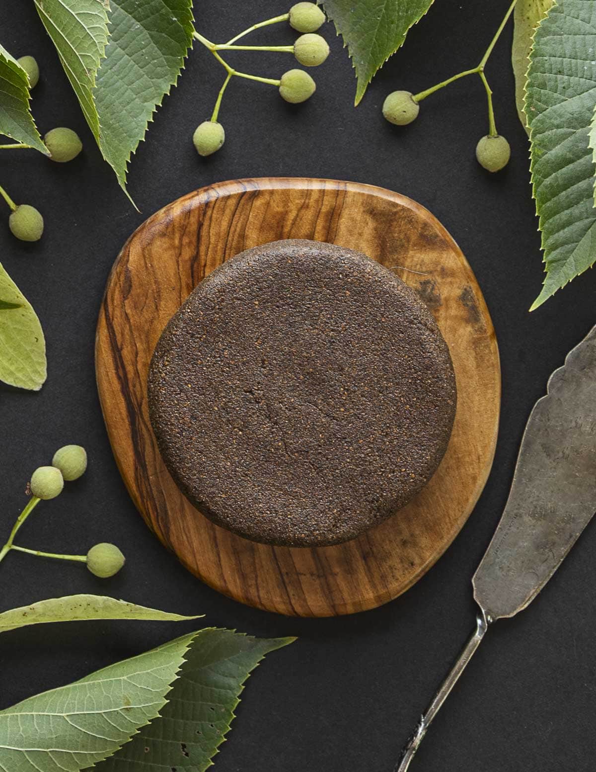 A round patty of roasted linden seed chocolate on a wooden olive wood board surrounded by basswood seeds.