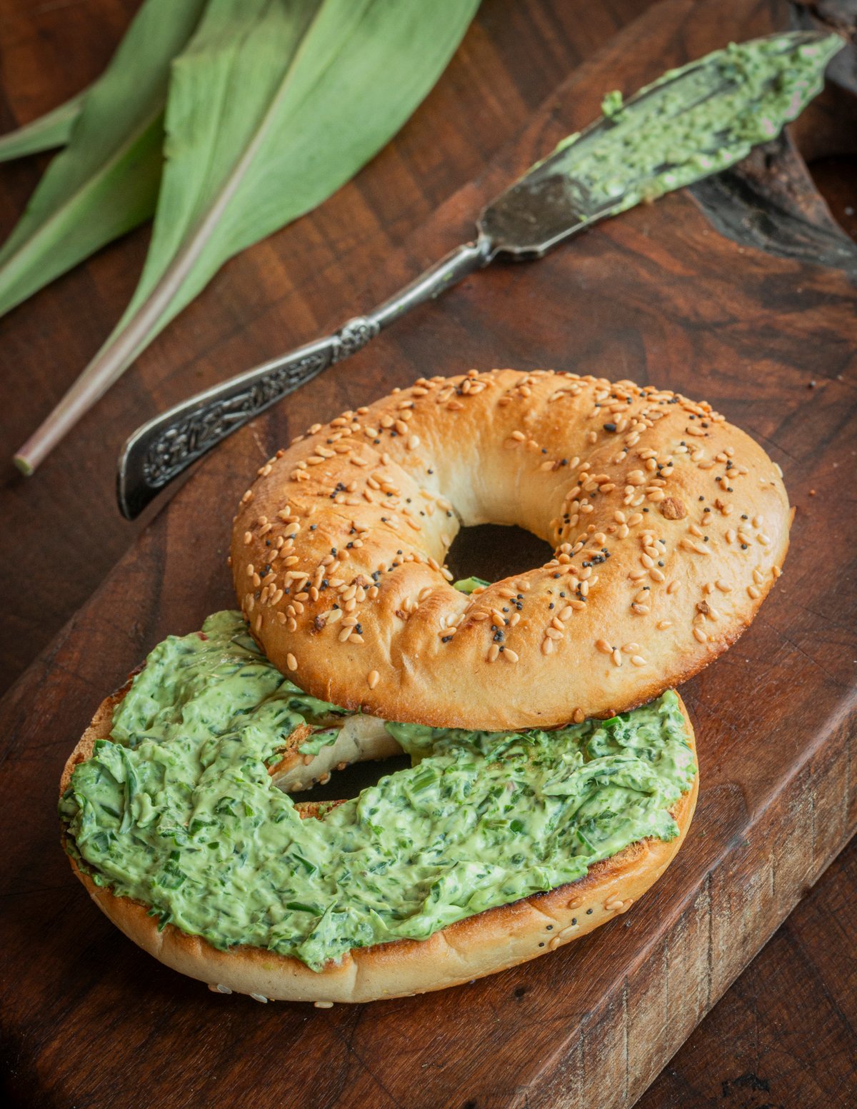 A bagel spread with wild ramp cream cheese with a knife and fresh ramp plants in the background.