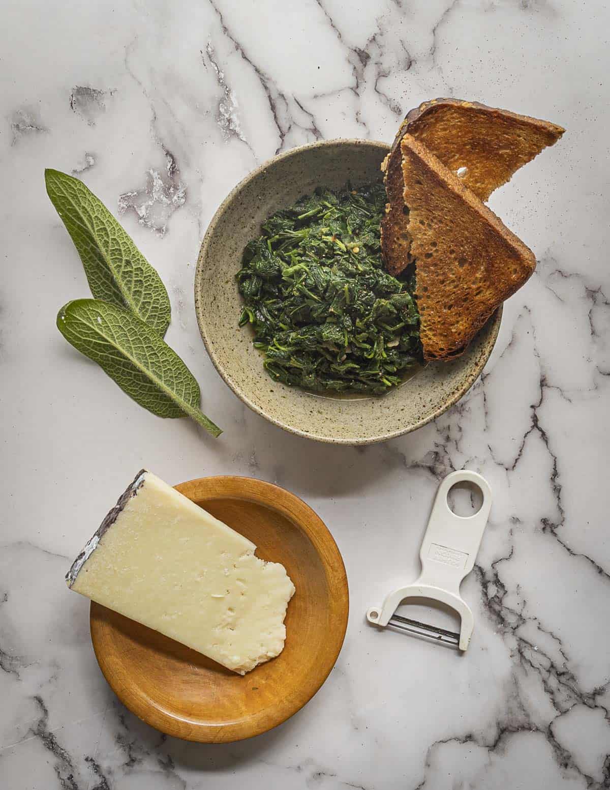 A bowl of cooked foraged greens including comfrey greens, garnished with grated pecorino cheese and served with bread. Fresh young comfrey leaves in the background. 