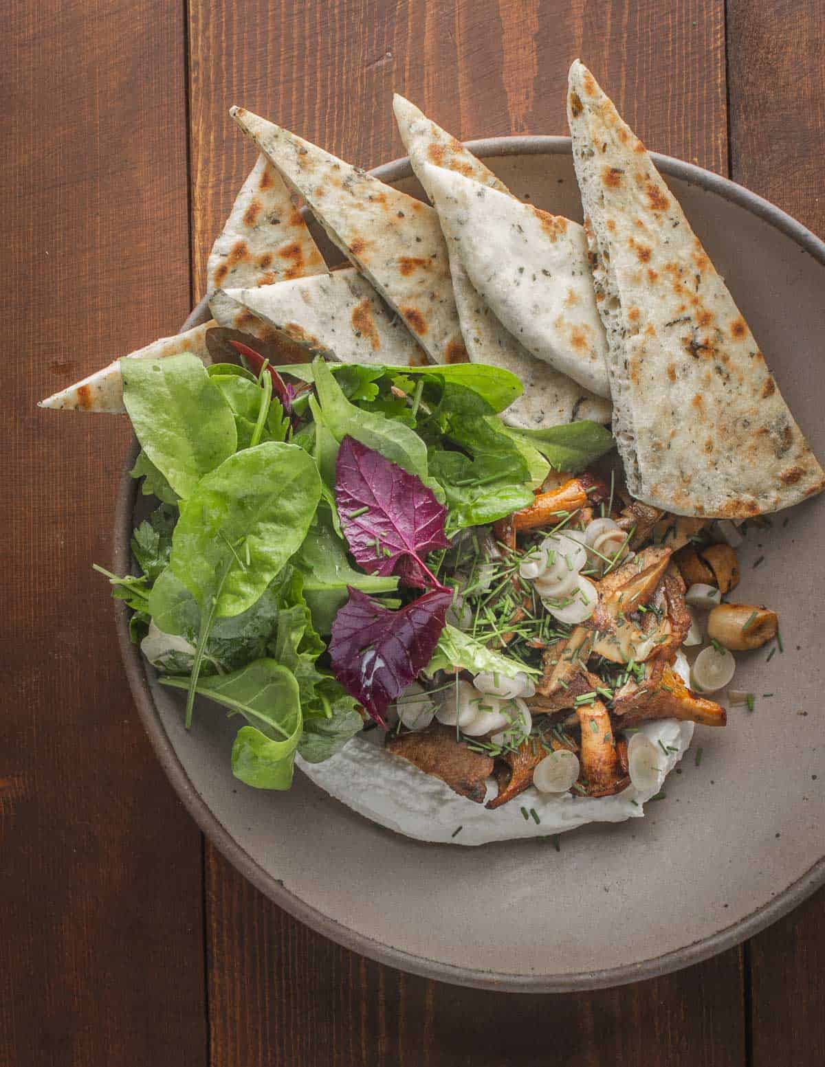 A bowl filled with wild herb flatbreads, whipped chevre, wild mushrooms and orache-nasturtium salad. 