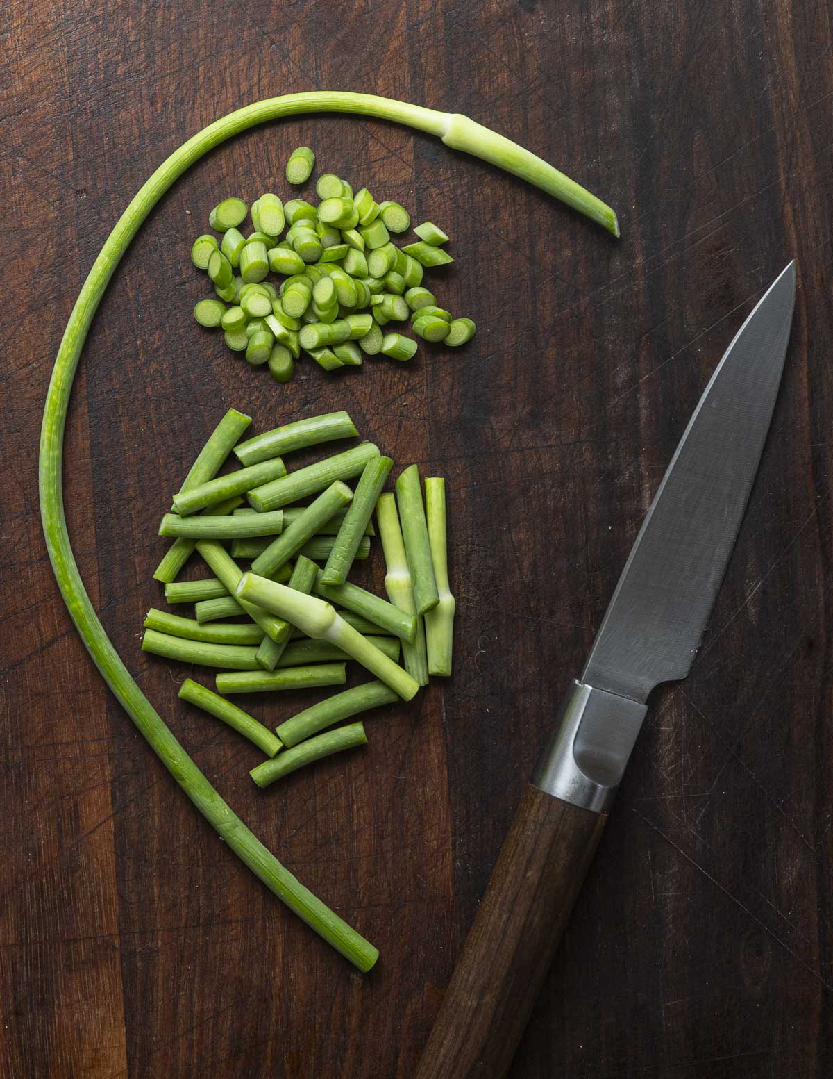 Garlic scapes cut into three different shapes on a cutting board showing the variety of ways they can be cut for cooking.
