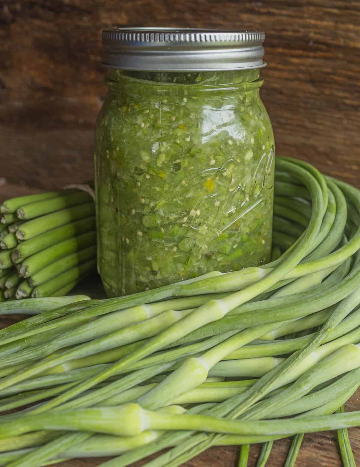 A jar of tomatillo and garlic scape salsa. surrounded by fresh garlic scapes.