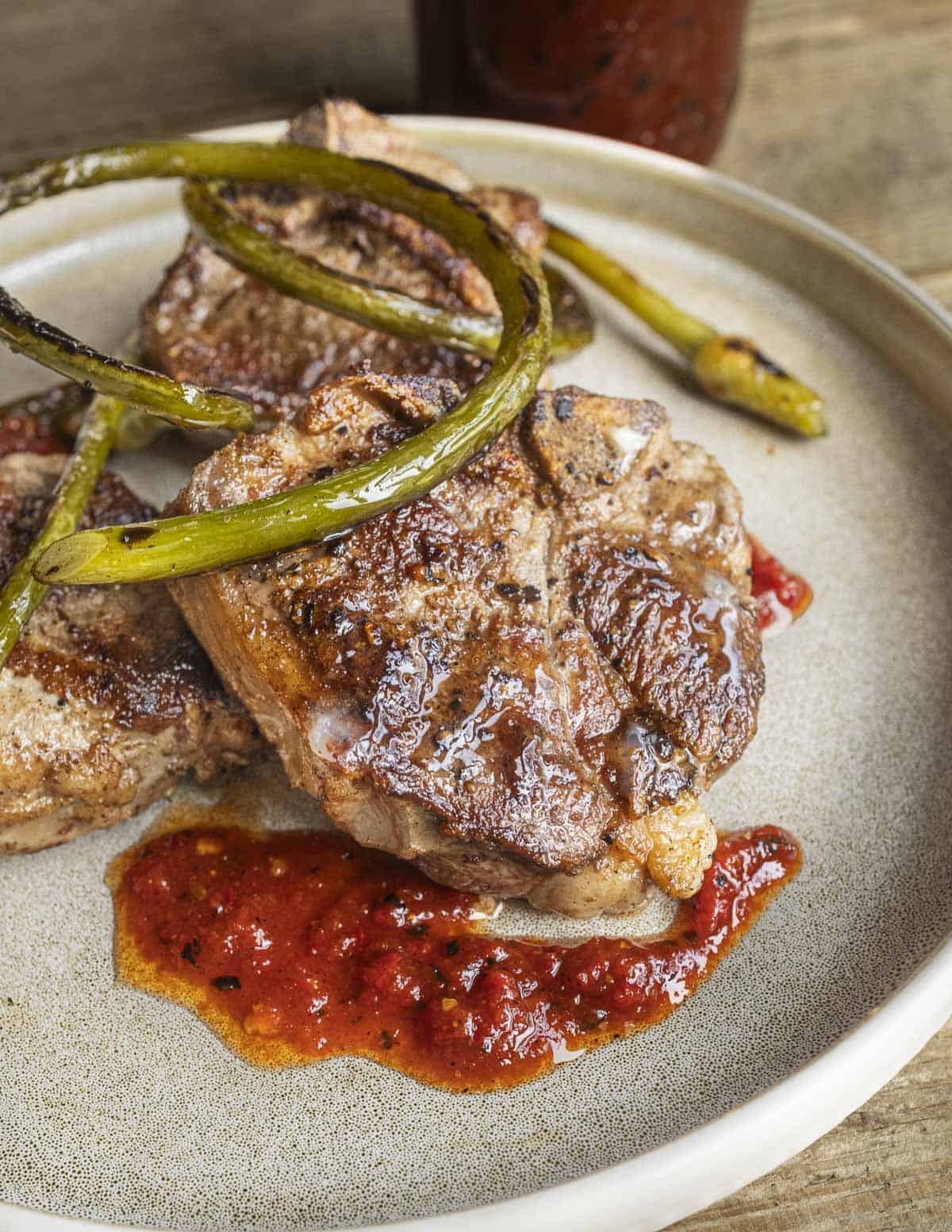 Lamb chops with a maple chili sauce on a plate garnished with grilled garlic scapes.