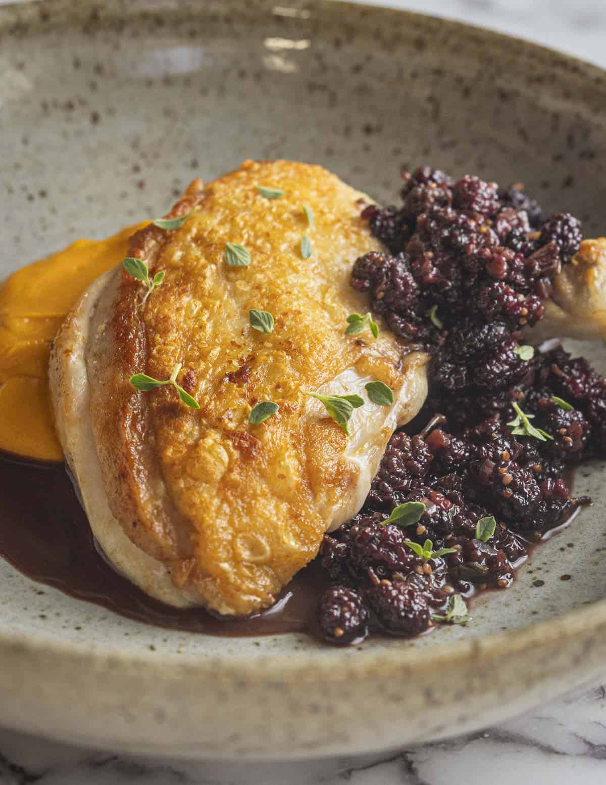 A pan roasted chicken breast with golden brown skin on a plate with carrot puree and mulberry sauce. 