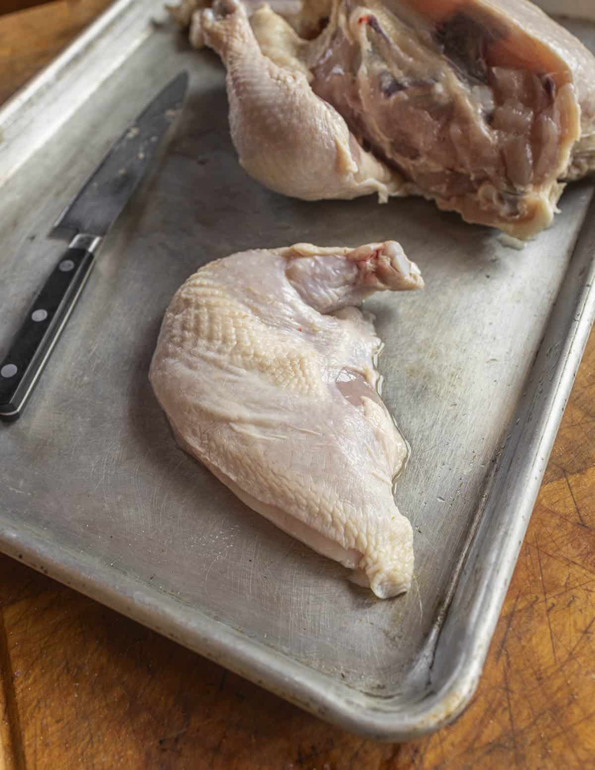 A bone-in, skin on or airline chicken breast on a baking sheet next to a knife and the chicken carcass that the breast was cut from. 