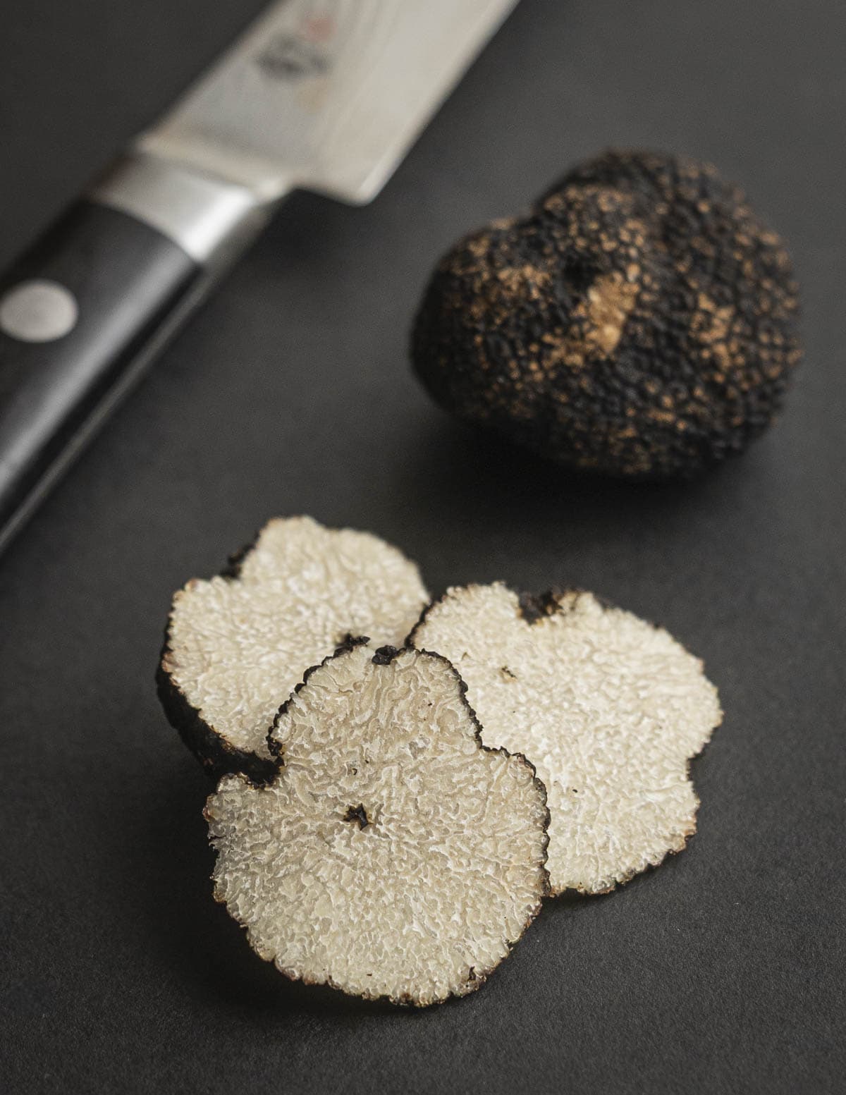 Sliced summer truffles showing a bright cream interior next to a whole truffle and a knife on a black background. 