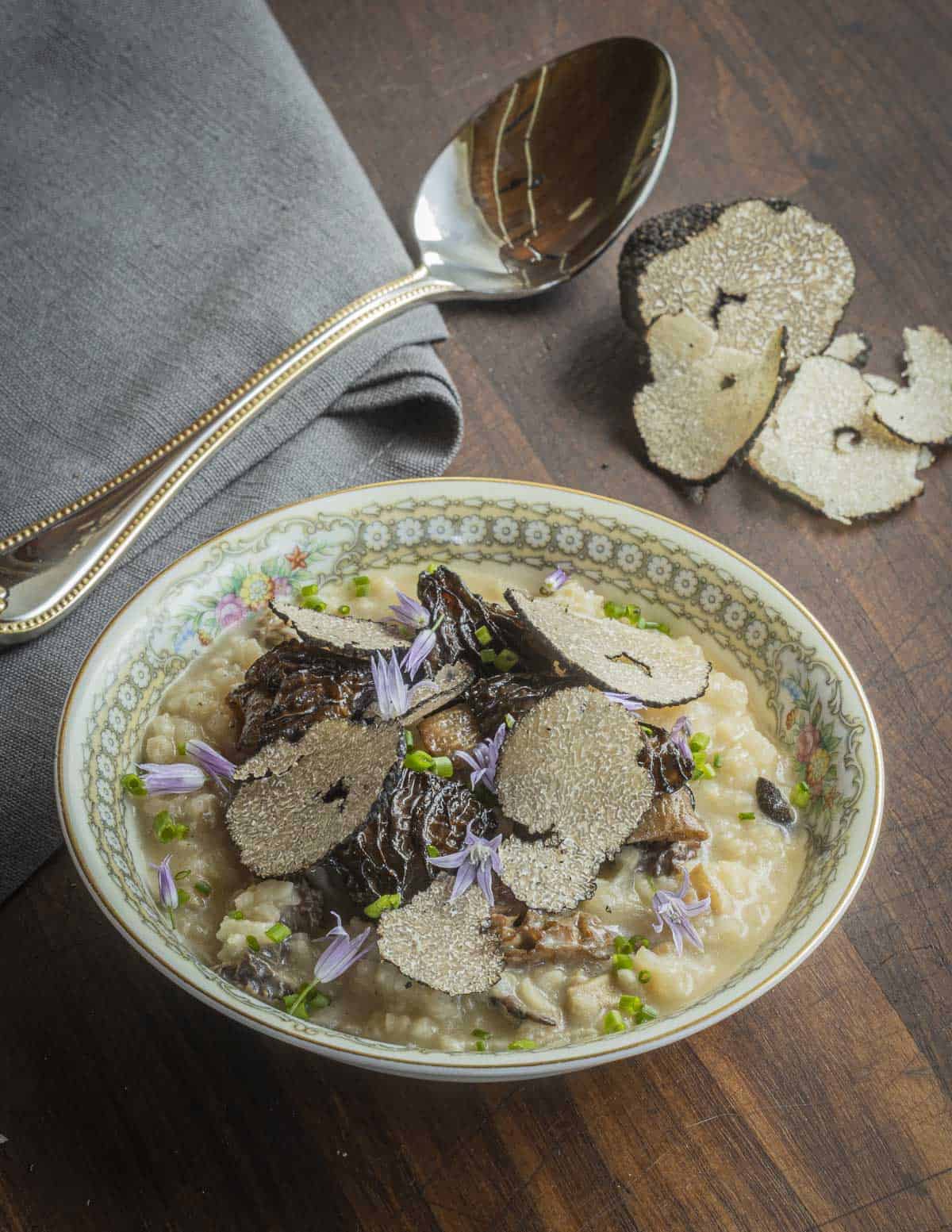 A bowl of risotto topped with morel mushrooms and shaved summer truffles with a cut truffle in the background. 