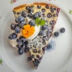 A perfectly cooked wild blueberry clafoutis on a plate garnished with whipped cream, marigold flowers, fresh blueberries, powdered sugar and mint leaves.