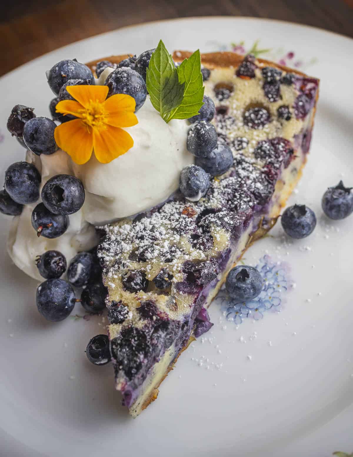 A slice of blueberry clafoutis dessert garnished with whipped cream wild blueberries and marigold flowers. 