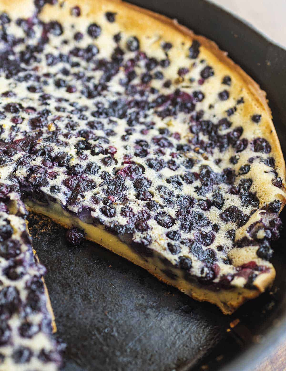 A finished blueberry clafoutis in a cast iron pan with a slice removed. 