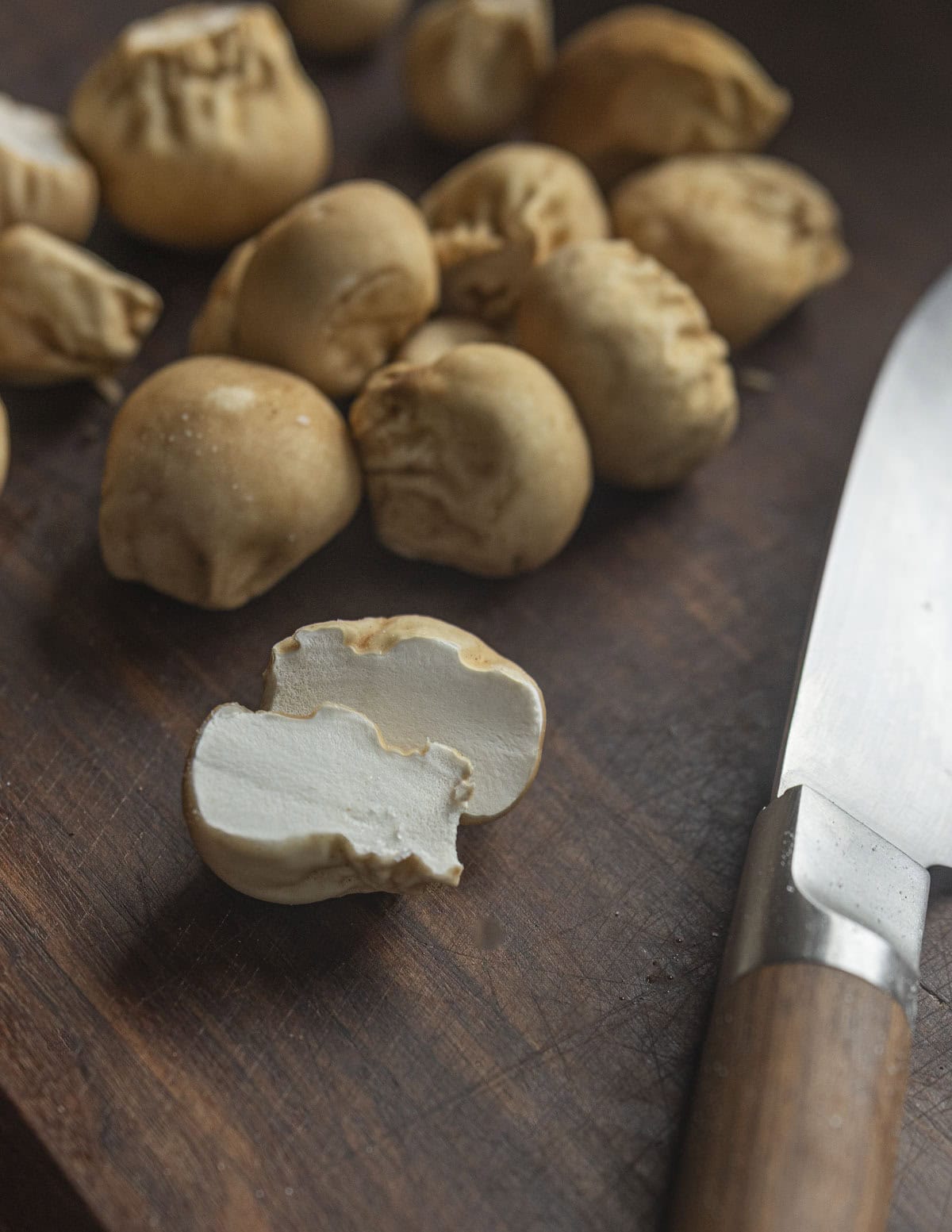 Numerous small brown puffball mushrooms on a cutting board next to a paring knife with a small pure white puffball cut in half in the foreground showing a perfect white interior. 
