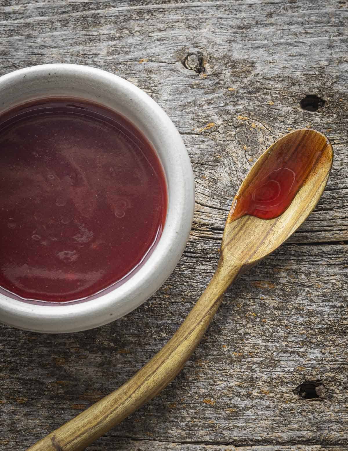 A red, shiny sweet and sour sauce made from highbush cranberry juice on a spoon carved from sumac wood next to a bowl of sauce.