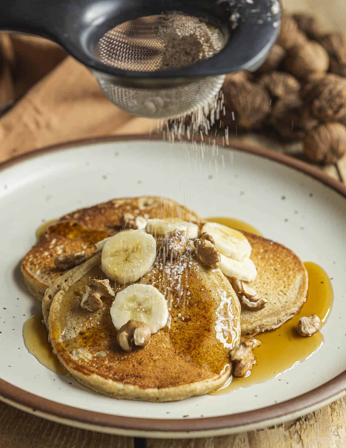 A hand holding a sieve dusting a plate of pancakes with spiced powdered sugar. 