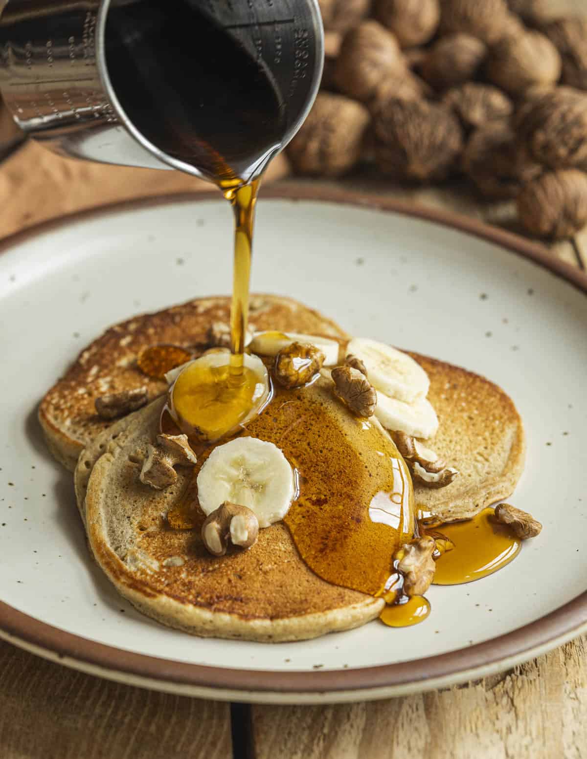 Pouring maple syrup on a pile of hot pancakes with bananas and walnuts.