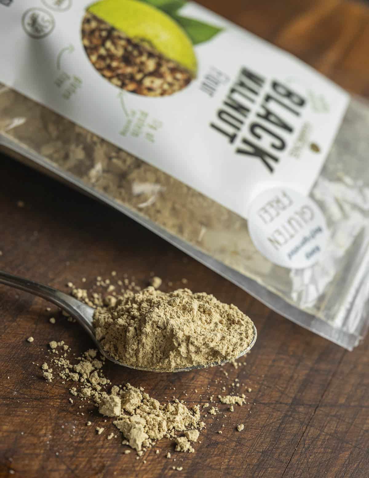 A spoon with black walnut flour spilling onto a cutting board next to a bag of walnut flour. 