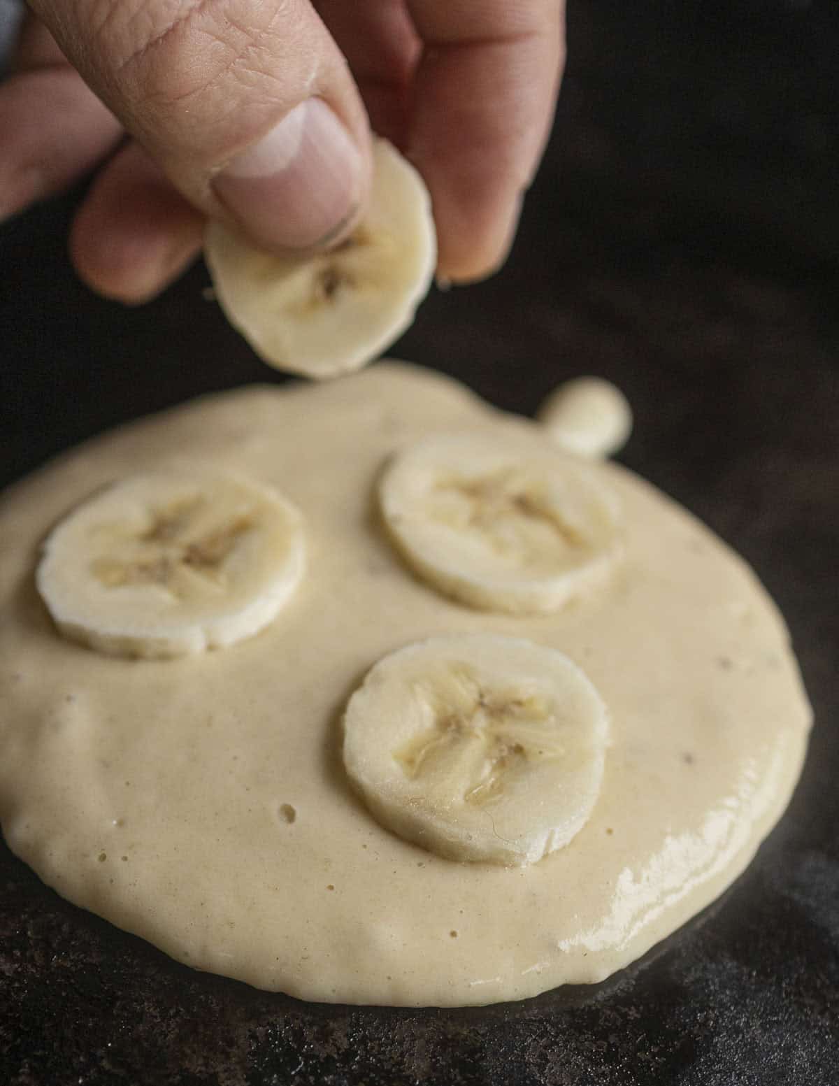 A hand laying a few sliced bananas on a pancake as it cooks. 