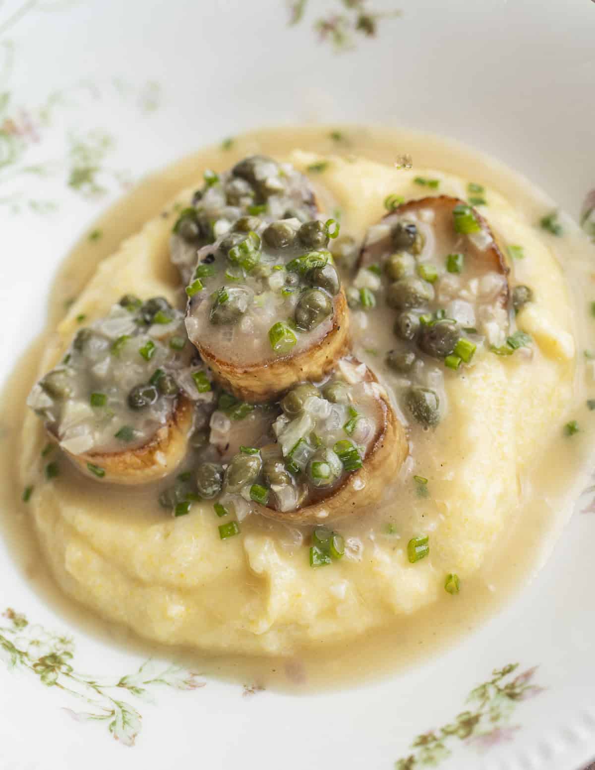 A finished plated dish of King oyster mushroom scallops on a bed of cooked yellow corn polenta with lemon, caper sauce and chives