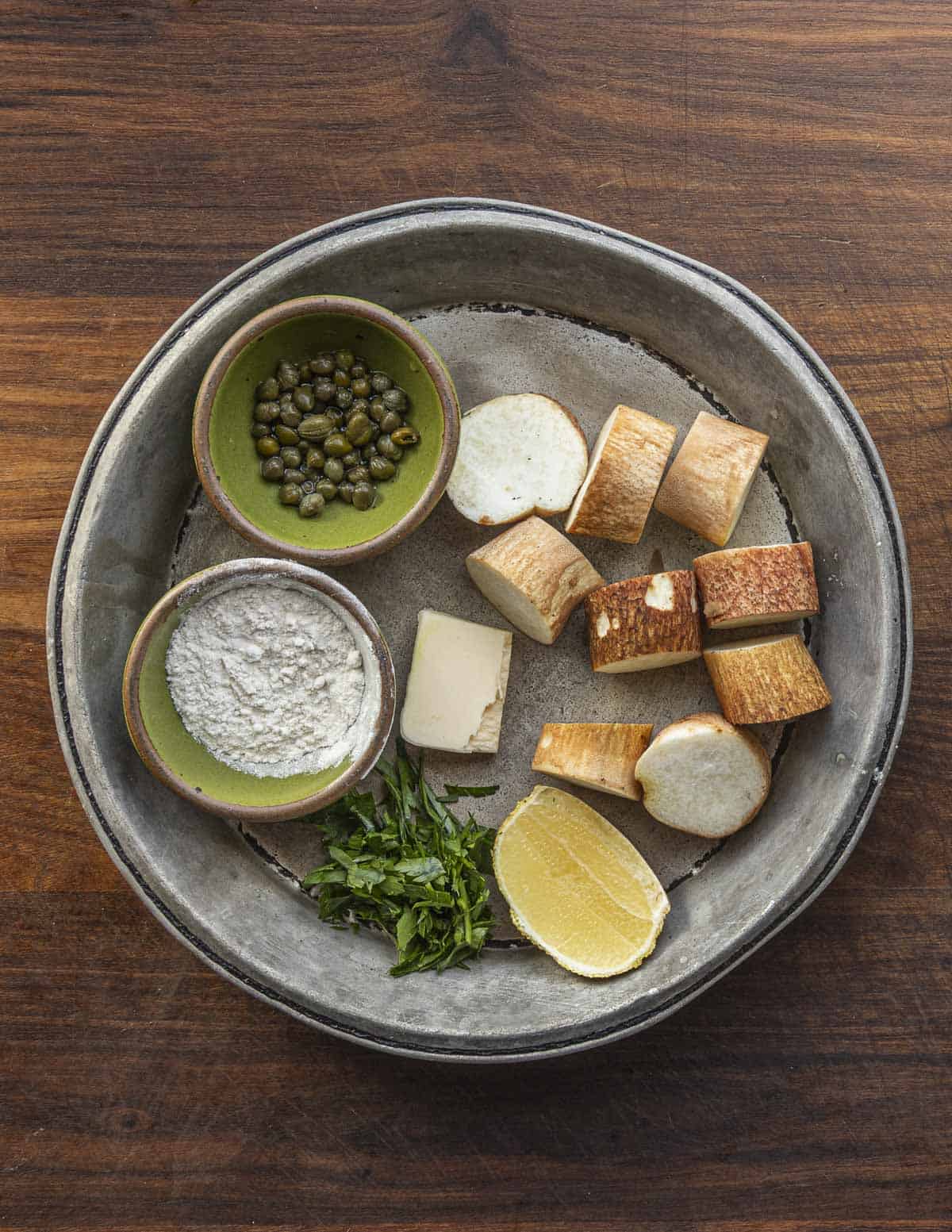 A top-down image of ingredients to make a Mushroom scallop recipe with lemon caper sauce: butter, Mushroom stems, parsley, capers and lemon shown in a pie plate.