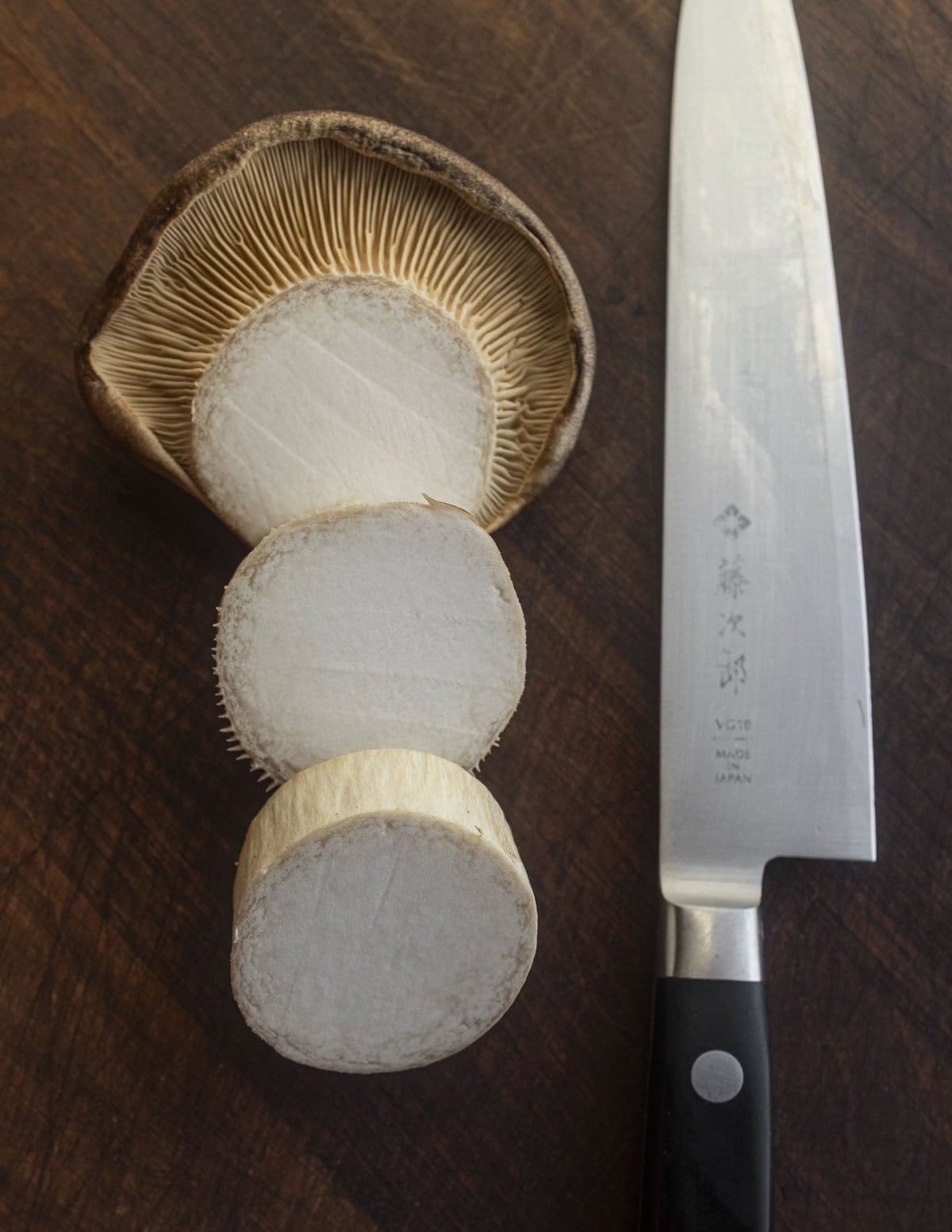 Thick slices of King oyster mushroom on a cutting board to be cooked as scallops next to a Japanese knife.