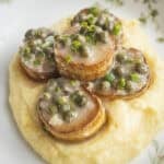 A close-up image of sautéed king oyster mushroom scallops on a bit of polenta with lemon, caper sauce, shallots, and herbs.