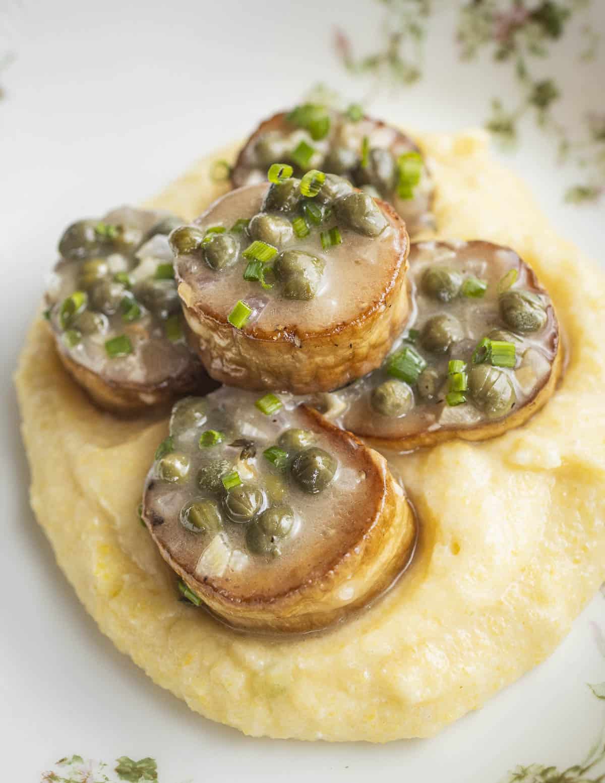 A close-up image of sautéed king oyster mushroom scallops on a bit of polenta with lemon, caper sauce, shallots, and herbs.